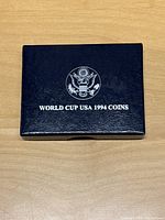 Closed navy blue presentation box with silver embossed US eagle and text 'WORLD CUP USA 1994 COINS' on lid, resting on wood surface.