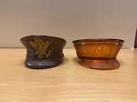 Two WWII soldier hat-shaped dresser containers, one glass amber with lid and one brown glazed clay without lid.
