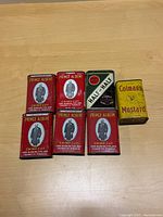 Seven rectangular metal tins from the early 1900s arranged on a wooden surface: five red Prince Albert Crimp Cut tobacco tins, one green and white Half and Half Burley and Bright tobacco tin, one yellow Colman's Mustard tin.