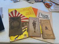 Photo shows five military and firearm reference booklets and brochures arranged on a cloth-covered surface.