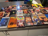 Photo of 25 assorted VHS movies laid out on a table with visible titles including Terminator, Pulp Fiction, Slap Shot, Reservoir Dogs, and Benny Hill collections.