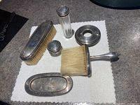 Full view of grooming set on white cloth, showing brushes, trays, containers, and glass bottle with silver lid.