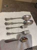Five pieces of sterling silverware on a towel including two large spoons, two forks, and one small spoon with ornate handles.