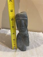 Front view of the stone carving showing one stylized face with carved eyes, nose, and lips next to measuring tape indicating approx 6.5 inches height.