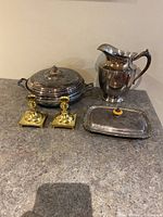Full view of silver plated serving dish, pitcher, small tray, and brass candlestick holders on countertop.
