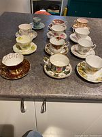 Ten assorted vintage fine bone china teacups and saucers arranged on a countertop showing variety of floral and gilded designs.