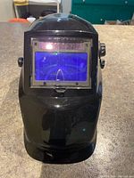 Front view of black welding helmet showing blue auto-darkening lens on the front.