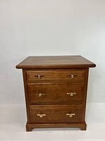 Front view of the wooden chest of drawers showing three drawers, brass handles, and keyholes. The wood has a polished finish and the chest rests on a slightly recessed base.