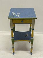 Front angle view of the hand painted one drawer stand showing the painted floral designs on blue surfaces and multi-colored legs.