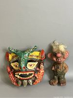 Photo showing both the Norwegian Nyform troll figurine and the tribal ceremonial pottery mask side by side on a gray surface.