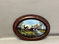 Front view of the oval vintage reverse painting on glass depicting a rural landscape with a house, stone bridge, river, and trees framed in a wood-tone oval frame.