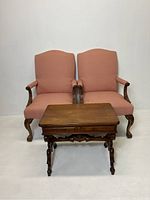 Pair of salmon upholstered armchairs with carved wooden arms and legs, positioned behind wooden end table with carved details and drawer