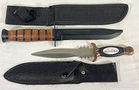 Both hunting knives laid side by side with their sheaths, showing the longer knife with stacked leather handle and black sheath, and the shorter knife with decorative handle and black leather sheath.