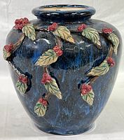 Front view of blue glazed pottery vase with dimensional green leaves and red berries decoration on one side.