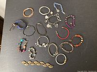 Photo showing 14 diverse bracelets including metal charm bracelet, beaded, braided leather, and decorative link designs, laid out on a black surface.