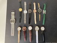 Full overhead view showing all 11 wristwatches arranged on a grey surface, displaying different strap colors and styles.