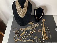 Photo of collection of various gold tone costume jewelry items displayed on black backdrop, including necklaces, bracelets, earrings, and pins.