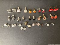 Full view of 24 pairs of men's cufflinks arranged in rows on a white surface, showing various styles and colors including silver tone, red, orange, black, and mother-of-pearl.