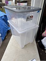 Two clear plastic storage totes stacked, top tote with grey lid and bottom tote with white lid, placed on a white table outdoors.