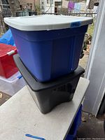 Two plastic storage totes stacked outside, one blue with a white lid on top of one black with a black lid.