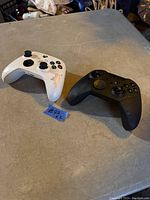 Top front view of two wireless Xbox Elite controllers, one white and one black, showing front buttons and thumbsticks with indication of wear on white controller