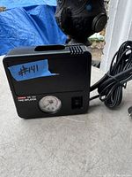 Photo of a black KOBER DC 12V tire inflator on a gray surface showing the pressure gauge, power switch, and attached power cord.