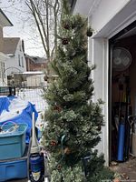 Full view of a 7' 3" tall artificial Christmas tree with green pine needles, pine cones, and string lights inside a garage doorway.