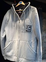 Light grey Ecko hoodie hanging on wooden hanger showing front side with zip zipper, hood with drawstrings, chest pocket with Ecko logo patch, and two hand pockets.
