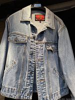 Front view of light blue denim jacket with button closures and pockets hanging on hanger.