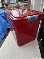 Image of large red plastic storage tote with lid placed on a white surface outdoors near a window with a backyard view.