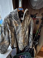Front view of men's camouflage winter jacket hanging on a wooden hanger, showing hood and front pockets.