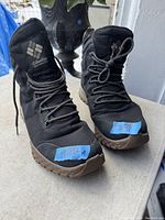 Front view of the men's black Columbia boots showing the brand logo on the tongue, size 11 labels on blue tape attached to toes, and black laces.