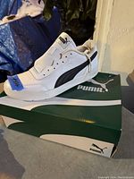 Left side view of one white Puma running shoe on top of green Puma box, showing black Puma logo stripe and white sole.