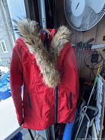 Red men's Rudsak winter jacket hanging on a hanger near a shelf. Shows front view with fur-lined hood and two zippered pockets.