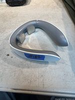 Photo showing light gray U-shaped neck massager with two metal electrode pads, placed on a white surface.