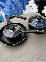 Photo showing two black Kichly saucepans with matching glass lids placed on top of the Lagostina frying pan. Handles are black with metal accents, lids have black knobs.