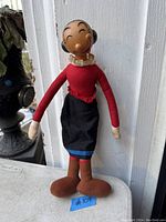 Vintage Olive Oyl doll standing against a white wooden background showing front view, dressed in red top, black skirt with blue trim, and brown shoes.