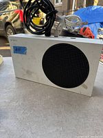 White Xbox Series S console showing black circular vent and two cables (power and HDMI) resting on surface in daylight.