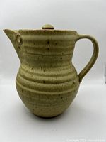 Front side view of antique stoneware pitcher showing its shape, handle and spout, with beige speckled glaze and concentric ridges.