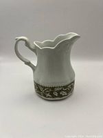 Side profile photo of the white ironstone creamer showing scalloped rim and olive green decorative floral border near the base.