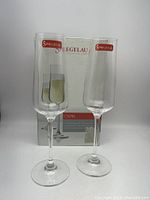 Two Spiegelau Capri champagne flutes with red label stickers on the rim, standing in front of their original box.