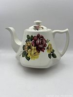 Front view of white ceramic tea pot with floral patterns and gold trimming on spout tip and lid knob, showing ornate handle and spout.