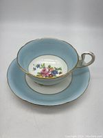 Tea cup and matching saucer set, showing the light blue rim, floral center design, and gold trim on the cup's edge and handle.