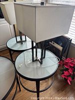 Pair of modern table lamps with rectangular white shades and geometric metal bases on round glass tops near window and red plant.