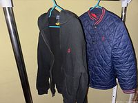 Two jackets hanging on rack: one black hooded zip-up and one navy blue quilted snap-button jacket with red logo
