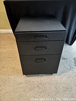 Front view of a black metal office cabinet with three closed drawers and caster wheels.