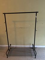 Front view of a bronze metal coat hanger with a bottom storage rack and hooks on the sides against a beige wall and floor.