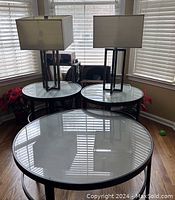 Photo showing three round tables arranged together in a corner with window blinds in background. Each table has a clear removable glass top and black metal frame. Two rectangular lamps sit on the end tables but are not included in the lot.