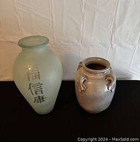 Two vases side by side on a black surface against a white background; left vase frosted glass with Asian characters, right vase gray ceramic with handles.