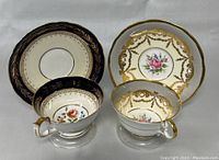 Front view of two vintage Aynsley teacups each paired with matching saucers. One set has navy blue band with gold leaf detailing, the other has a white base with gold ornate garland and pink rose floral pattern.